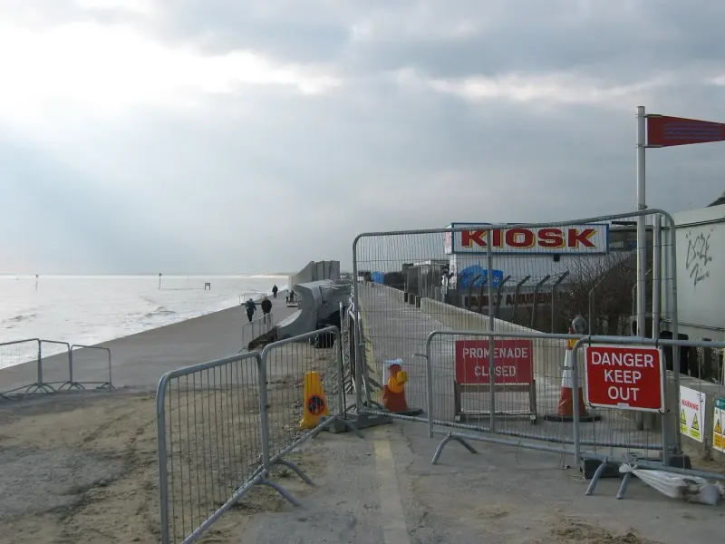 A closed seafront promenade behind site fencing, with the sea wall works beyond